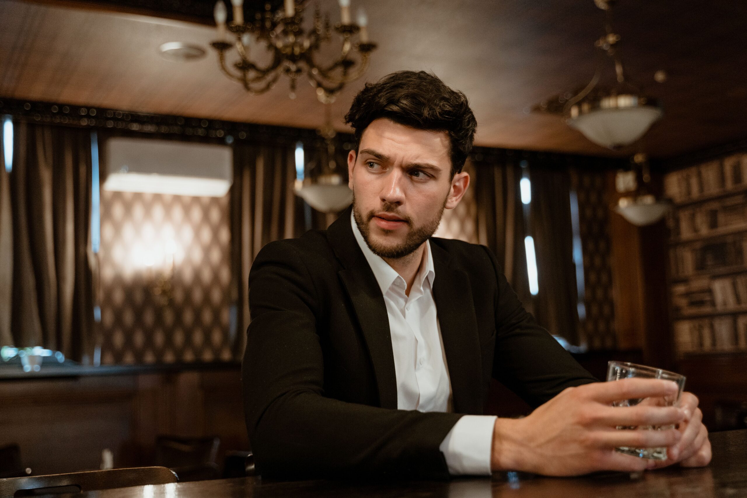A man in a black suit with a drink, sitting at a bar in a sophisticated setting.